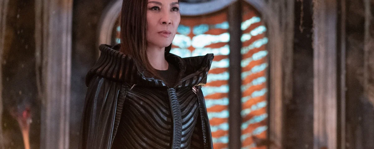 Une femme vêtue d'un costume noir détaillé aux épaules structurées se tient à l'intérieur, incarnant Michelle Yeoh, devant une fenêtre cintrée éclairée par des lumières horizontales orange et blanches.