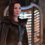 Une femme vêtue d'un costume noir détaillé aux épaules structurées se tient à l'intérieur, incarnant Michelle Yeoh, devant une fenêtre cintrée éclairée par des lumières horizontales orange et blanches.