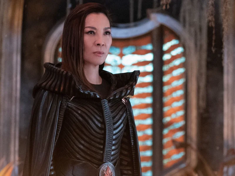 Une femme vêtue d'un costume noir détaillé aux épaules structurées se tient à l'intérieur, incarnant Michelle Yeoh, devant une fenêtre cintrée éclairée par des lumières horizontales orange et blanches.