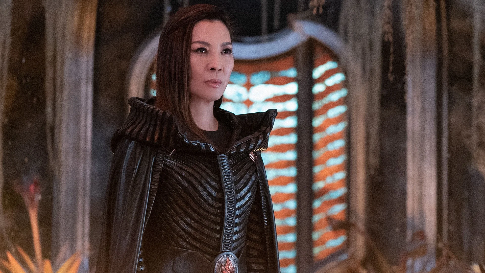 Une femme vêtue d'un costume noir détaillé aux épaules structurées se tient à l'intérieur, incarnant Michelle Yeoh, devant une fenêtre cintrée éclairée par des lumières horizontales orange et blanches.