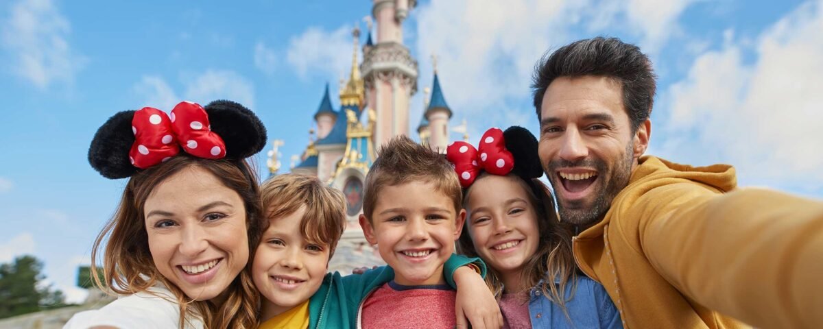 Une famille de cinq personnes sourit et pose pour un selfie avec ses enfants devant un château de conte de fées à Disneyland Paris par une journée ensoleillée.