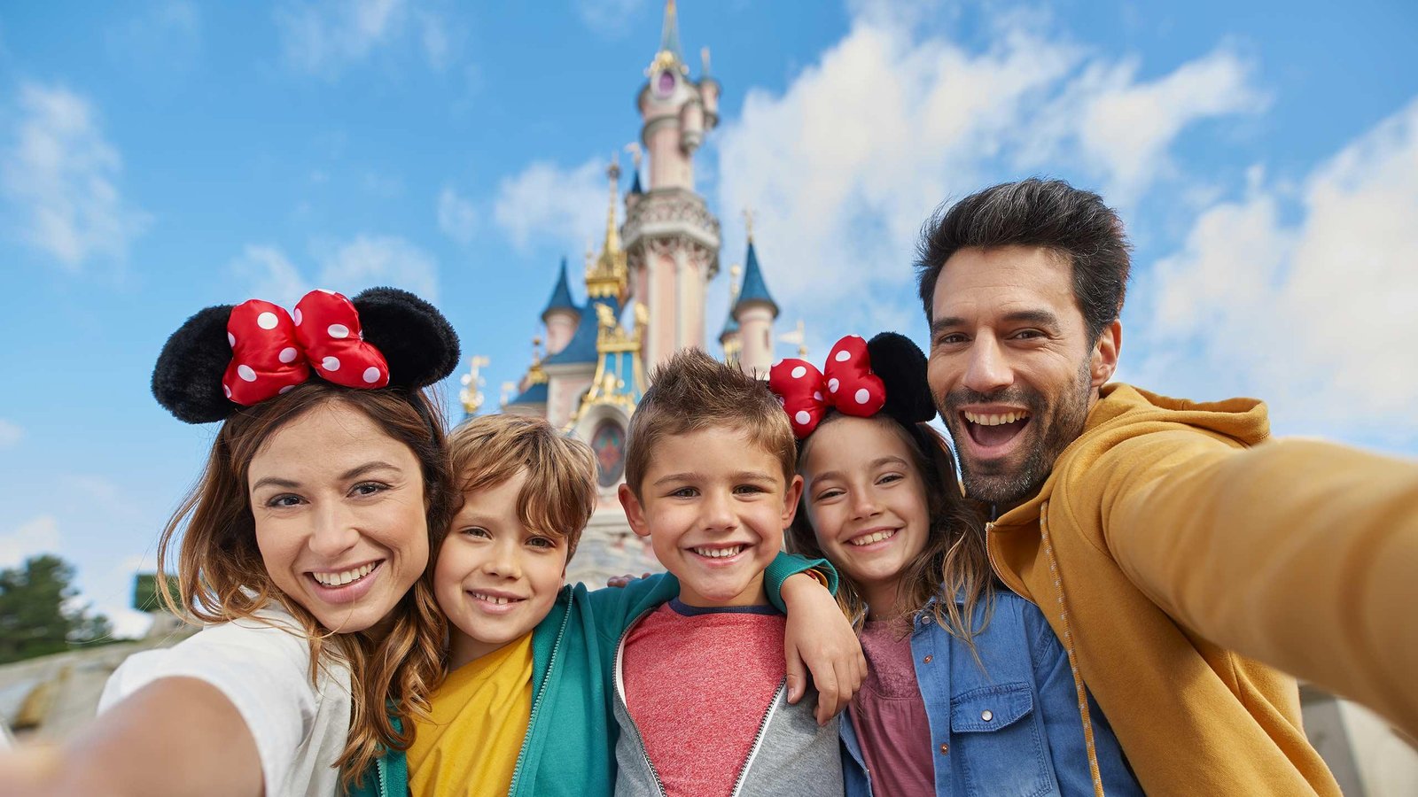 Une famille de cinq personnes sourit et pose pour un selfie avec ses enfants devant un château de conte de fées à Disneyland Paris par une journée ensoleillée.