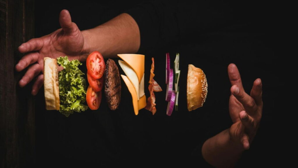 Un hamburger déconstruit avec des ingrédients flottants - pain, laitue, tomate, galette de bœuf, fromage, bacon, oignon, cornichons, pain supérieur - entre deux mains levées. Fait partie de l'édition limitée de la collection Les Burgers Avengers 2026.