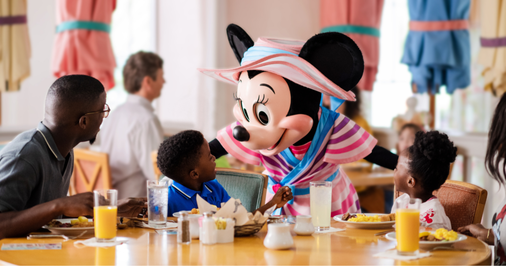 Une Minnie Mouse costumée accueille deux enfants à une table de restaurant pendant que les adultes regardent ; le petit déjeuner et les boissons sont sur la table - parfait pour les familles qui profitent du Free Dining Disney ou qui explorent les offres du Plan Repas Hôtels Disney.
