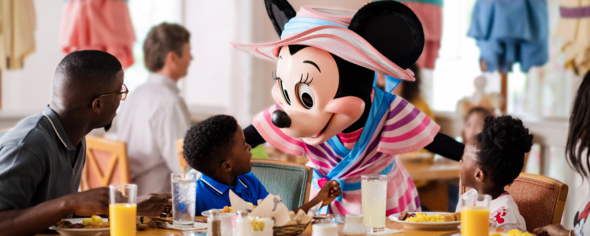 Une Minnie Mouse costumée accueille deux enfants à une table de restaurant pendant que les adultes regardent ; le petit déjeuner et les boissons sont sur la table - parfait pour les familles qui profitent du Free Dining Disney ou qui explorent les offres du Plan Repas Hôtels Disney.