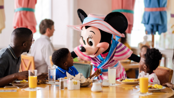 Une Minnie Mouse costumée accueille deux enfants à une table de restaurant pendant que les adultes regardent ; le petit déjeuner et les boissons sont sur la table - parfait pour les familles qui profitent du Free Dining Disney ou qui explorent les offres du Plan Repas Hôtels Disney.