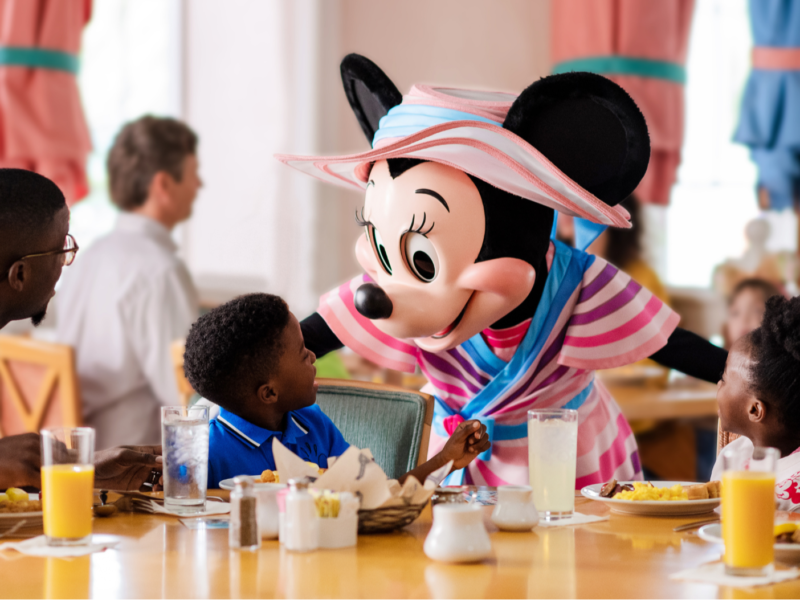 Une Minnie Mouse costumée accueille deux enfants à une table de restaurant pendant que les adultes regardent ; le petit déjeuner et les boissons sont sur la table - parfait pour les familles qui profitent du Free Dining Disney ou qui explorent les offres du Plan Repas Hôtels Disney.