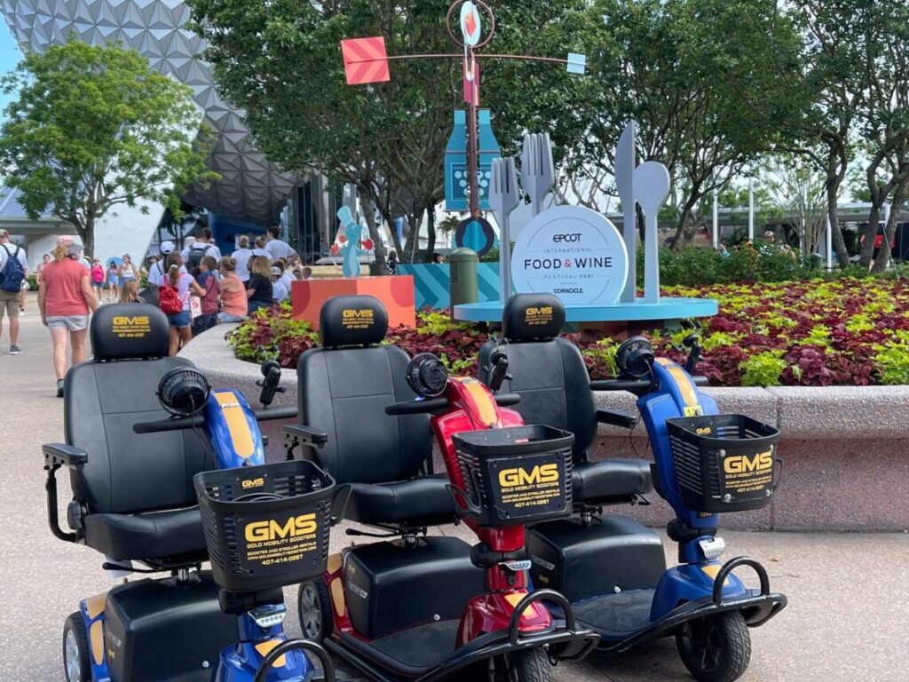 Trois scooters électriques de location sont garés devant un panneau du EPCOT International Food & Wine Festival, avec des visiteurs et de la verdure en arrière-plan, rappelant les scènes festives de Disneyland Paris ou de Chalifert.
