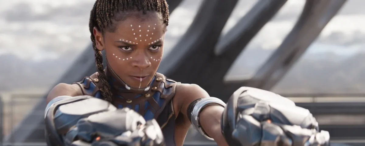 Une femme aux cheveux tressés, portant une armure futuriste et des gantelets métalliques comme Shuri Black Panther, se tient à l'extérieur, les bras étendus, l'air concentré.
