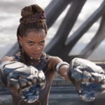 Une femme aux cheveux tressés, portant une armure futuriste et des gantelets métalliques comme Shuri Black Panther, se tient à l'extérieur, les bras étendus, l'air concentré.