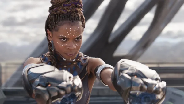 Une femme aux cheveux tressés, portant une armure futuriste et des gantelets métalliques comme Shuri Black Panther, se tient à l'extérieur, les bras étendus, l'air concentré.