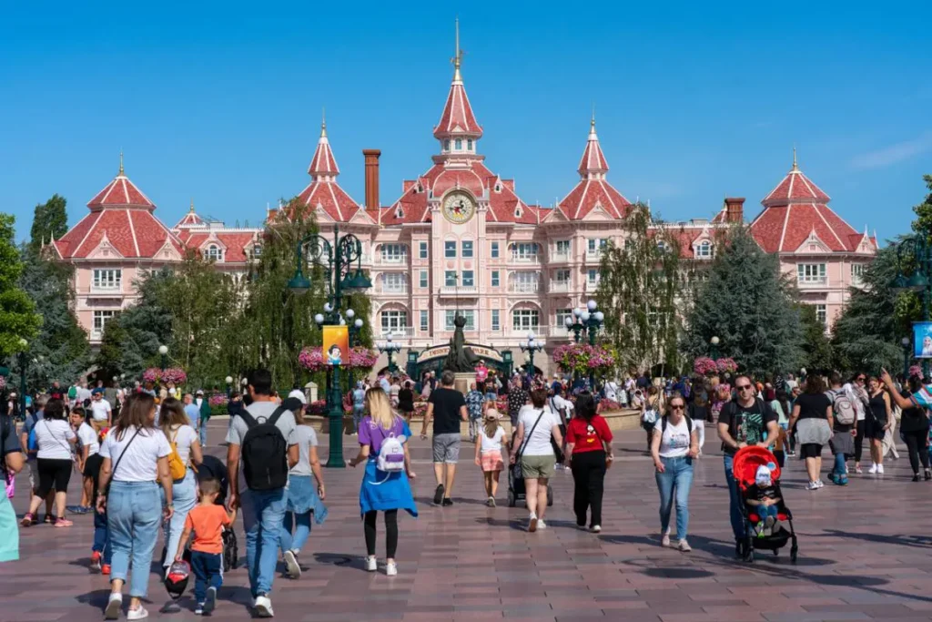 Une foule de personnes se dirige vers un grand bâtiment rose de style victorien avec des toits rouges et une horloge au-dessus de l'entrée, profitant de l'atmosphère accueillante et des services visiteurs Disney lors d'une journée ensoleillée dans les parcs Disney.