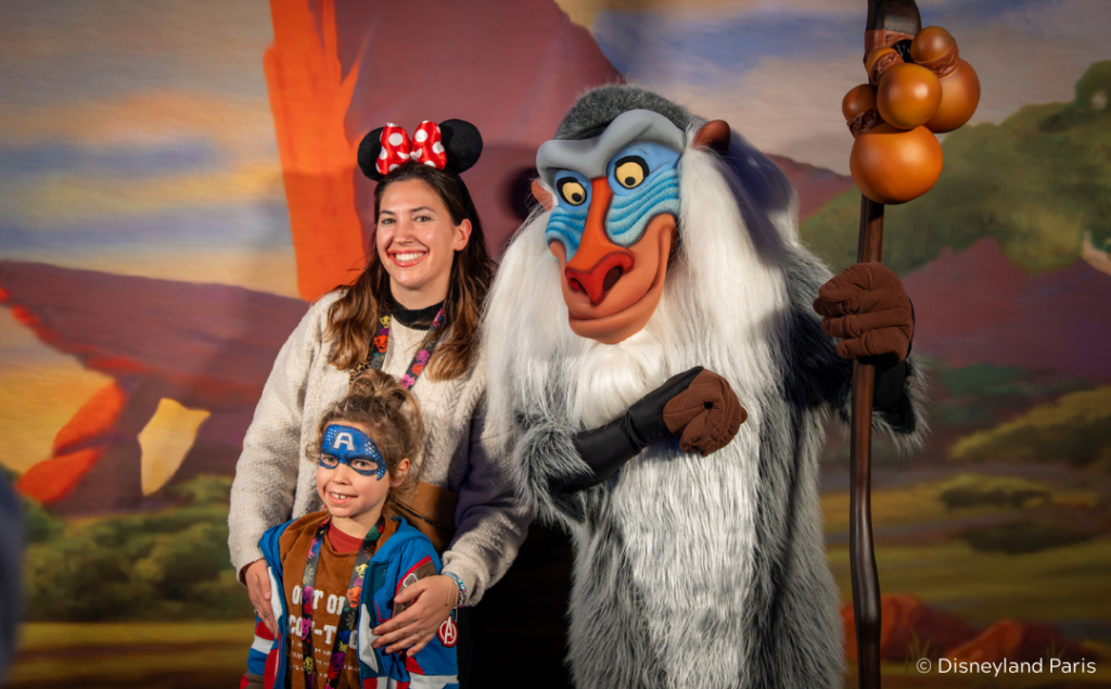 Une femme et un enfant posent avec Rafiki du Roi Lion devant une toile de fond à Disneyland Paris pour soutenir l'initiative Pièces Jaunes 2026 produits solidaires.