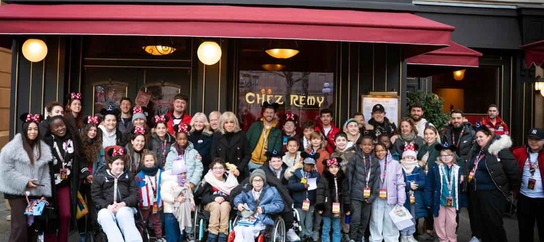 Un grand groupe d'enfants et d'adultes, certains en fauteuil roulant et beaucoup portant des oreilles de Mickey, posent pour une photo devant le restaurant Bistrot Chez Rémy à Disneyland Paris pendant l'initiative Pièces Jaunes 2026.