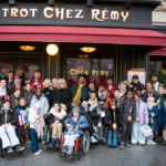 Un grand groupe d'enfants et d'adultes, certains en fauteuil roulant et beaucoup portant des oreilles de Mickey, posent pour une photo devant le restaurant Bistrot Chez Rémy à Disneyland Paris pendant l'initiative Pièces Jaunes 2026.