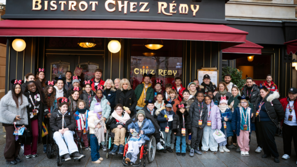 Un grand groupe d'enfants et d'adultes, certains en fauteuil roulant et beaucoup portant des oreilles de Mickey, posent pour une photo devant le restaurant Bistrot Chez Rémy à Disneyland Paris pendant l'initiative Pièces Jaunes 2026.