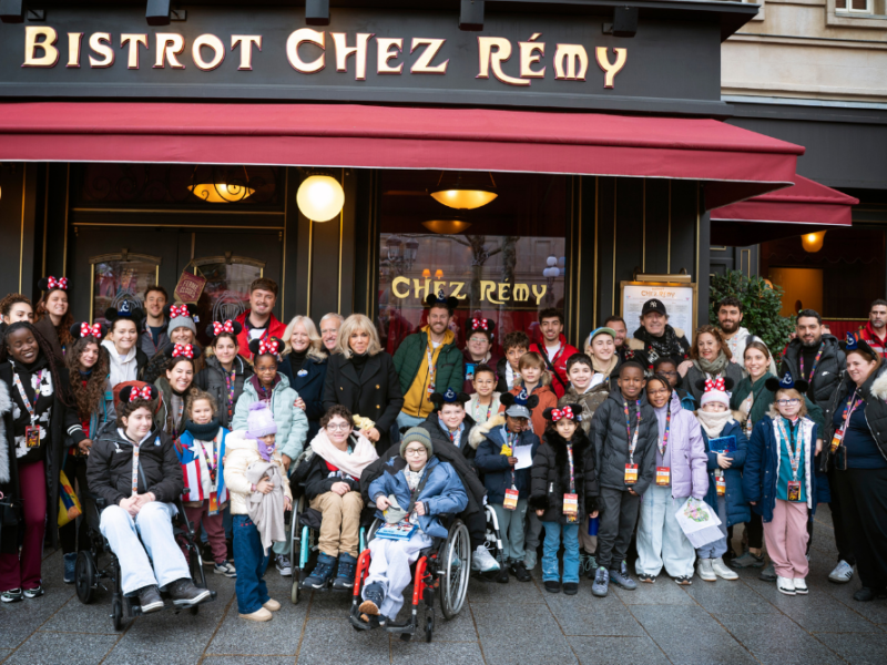 Un grand groupe d'enfants et d'adultes, certains en fauteuil roulant et beaucoup portant des oreilles de Mickey, posent pour une photo devant le restaurant Bistrot Chez Rémy à Disneyland Paris pendant l'initiative Pièces Jaunes 2026.