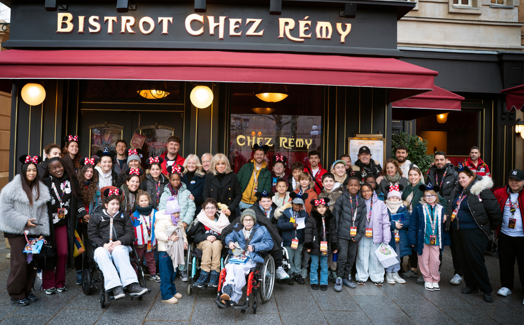Un grand groupe d'enfants et d'adultes, certains en fauteuil roulant et beaucoup portant des oreilles de Mickey, posent pour une photo devant le restaurant Bistrot Chez Rémy à Disneyland Paris pendant l'initiative Pièces Jaunes 2026.