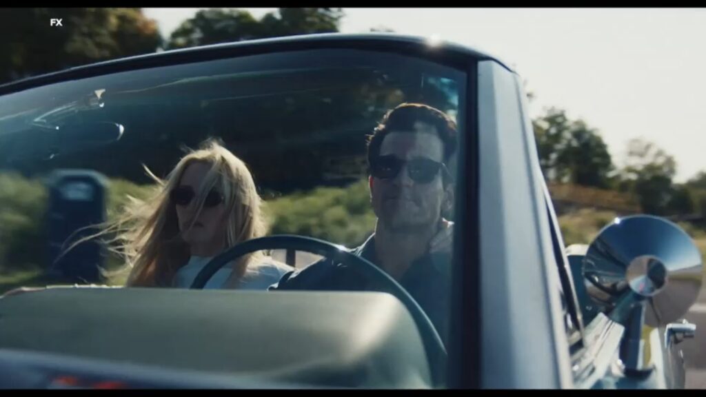 Un homme et une femme portant des lunettes de soleil conduisent une voiture décapotable avec le toit baissé par une journée ensoleillée, capturant le glamour d'un classique de Ryan Murphy FX Love Story.