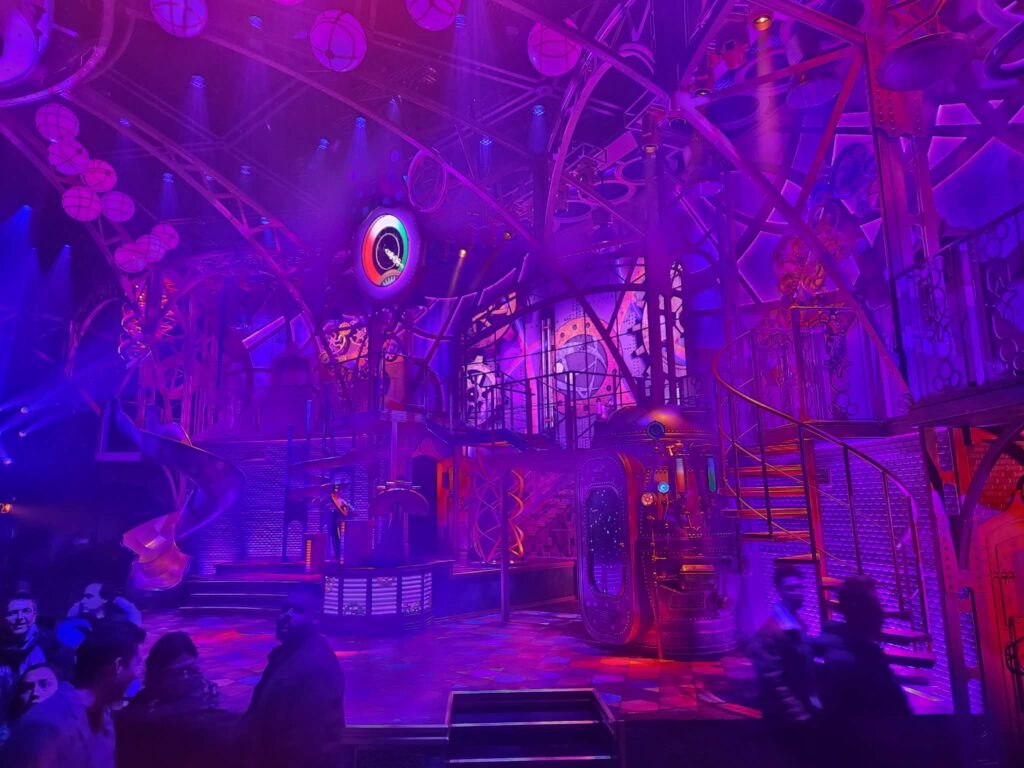 Des personnes se tiennent devant une scène futuriste très éclairée à Disneyland Paris, avec des lumières violettes et roses, des escaliers en colimaçon et un décor industriel, donnant vie à la magie de la Fabrique des Rêves.
