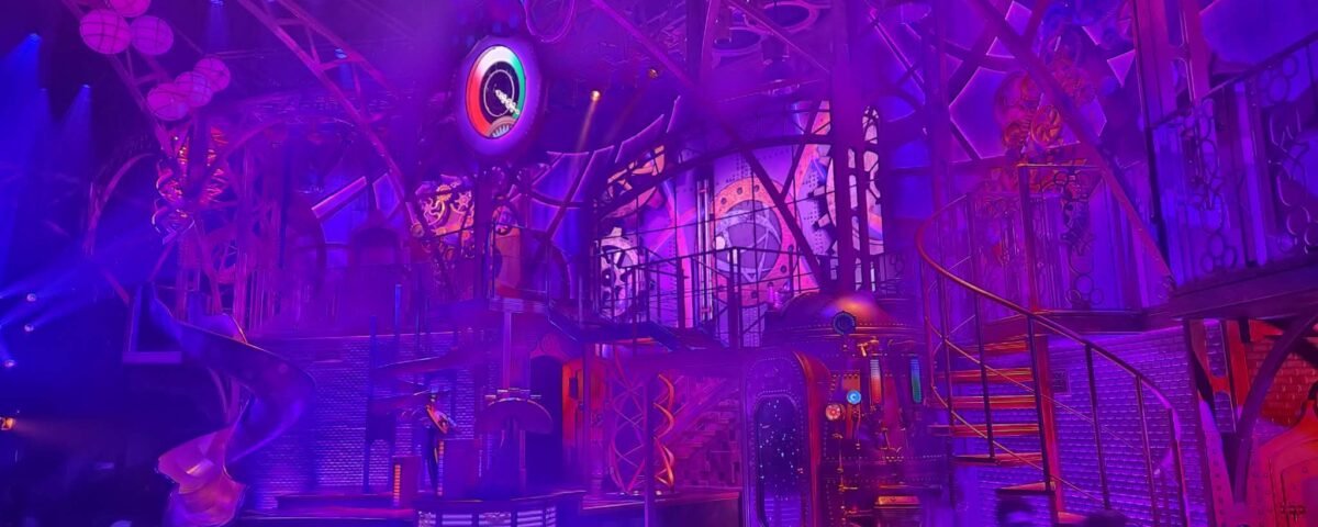 Des personnes se tiennent devant une scène futuriste très éclairée à Disneyland Paris, avec des lumières violettes et roses, des escaliers en colimaçon et un décor industriel, donnant vie à la magie de la Fabrique des Rêves.
