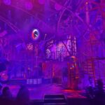 Des personnes se tiennent devant une scène futuriste très éclairée à Disneyland Paris, avec des lumières violettes et roses, des escaliers en colimaçon et un décor industriel, donnant vie à la magie de la Fabrique des Rêves.