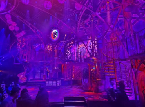 Des personnes se tiennent devant une scène futuriste très éclairée à Disneyland Paris, avec des lumières violettes et roses, des escaliers en colimaçon et un décor industriel, donnant vie à la magie de la Fabrique des Rêves.