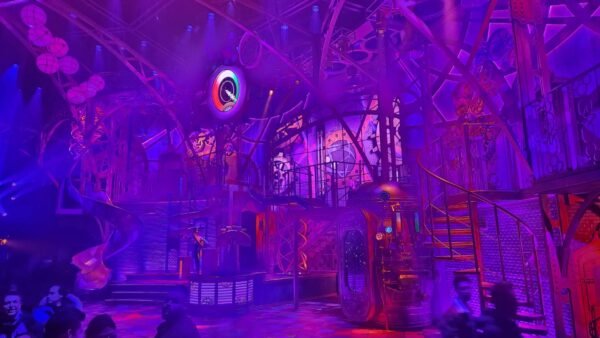 Des personnes se tiennent devant une scène futuriste très éclairée à Disneyland Paris, avec des lumières violettes et roses, des escaliers en colimaçon et un décor industriel, donnant vie à la magie de la Fabrique des Rêves.