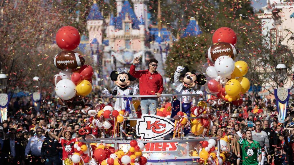 Une personne portant un maillot rouge des Kansas City Chiefs se tient sur un char de parade avec Mickey et Minnie Mouse à Disneyland, entourée de ballons et de confettis, célébrant une tradition du Super Bowl tandis qu'une foule applaudit en arrière-plan.