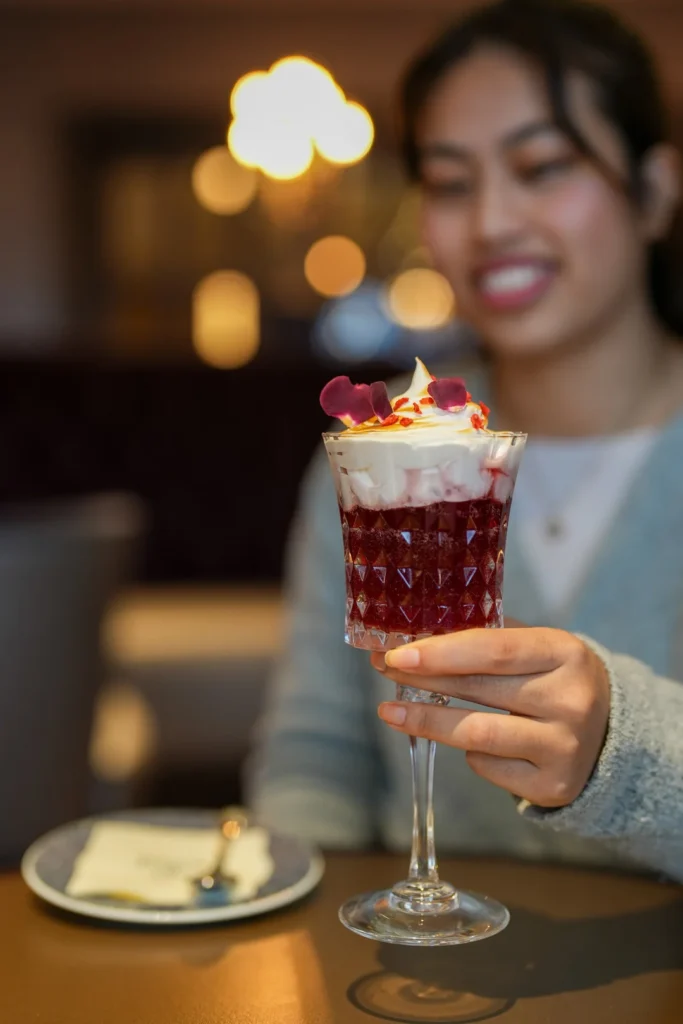 Une personne tient un verre décoratif rempli d'un dessert en couches garni de crème fouettée et de fleurs comestibles, idéal pour déguster des cocktails duo exclusifs à Disneyland Paris, avec une petite assiette et une cuillère sur la table.