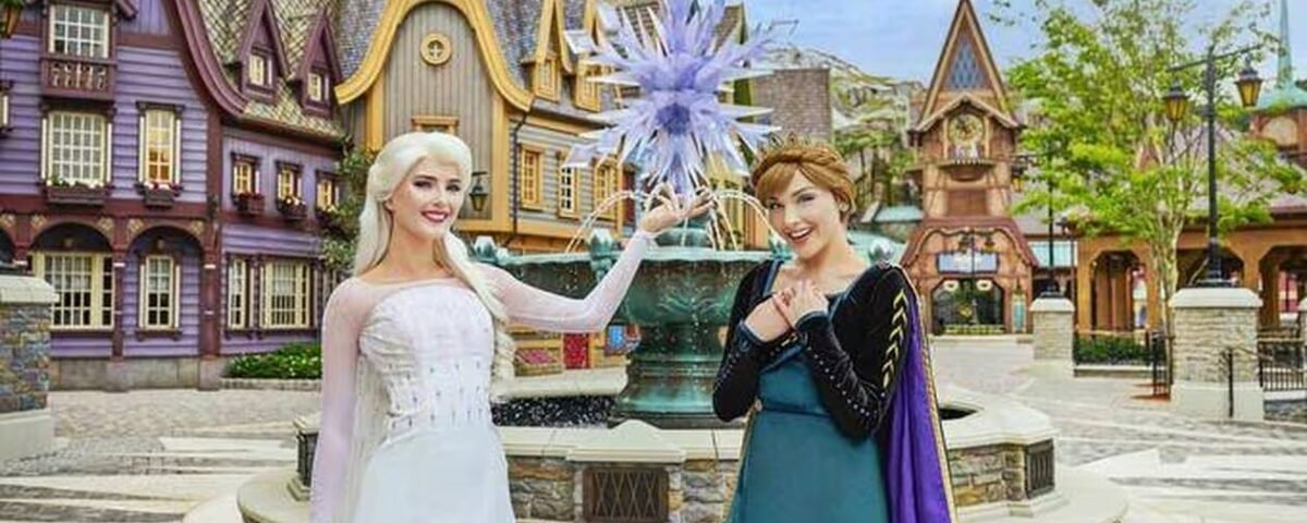 Deux femmes déguisées en Elsa et Anna de La Reine des Neiges posent devant une fontaine à thème et des bâtiments de style villageois, capturant la magie des attractions de Disneyland Paris.