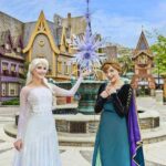 Deux femmes déguisées en Elsa et Anna de La Reine des Neiges posent devant une fontaine à thème et des bâtiments de style villageois, capturant la magie des attractions de Disneyland Paris.
