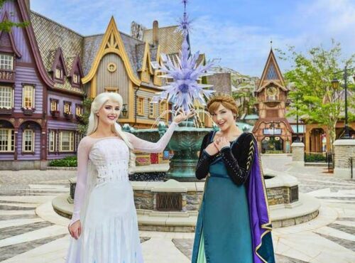 Deux femmes déguisées en Elsa et Anna de La Reine des Neiges posent devant une fontaine à thème et des bâtiments de style villageois, capturant la magie des attractions de Disneyland Paris.