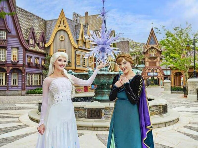 Deux femmes déguisées en Elsa et Anna de La Reine des Neiges posent devant une fontaine à thème et des bâtiments de style villageois, capturant la magie des attractions de Disneyland Paris.