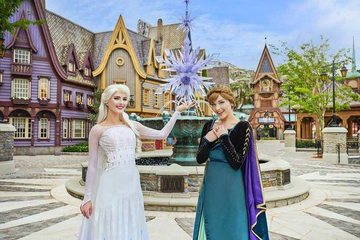Deux femmes déguisées en Elsa et Anna de La Reine des Neiges posent devant une fontaine à thème et des bâtiments de style villageois, capturant la magie des attractions de Disneyland Paris.