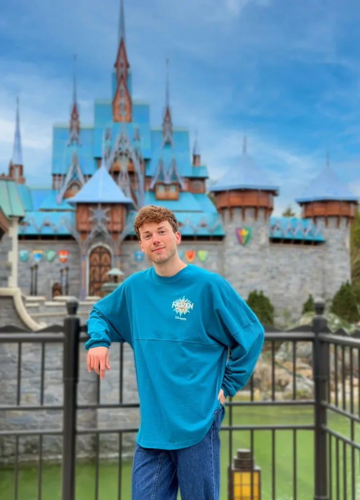 Un jeune homme vêtu d'un sweat-shirt bleu sur le thème de Frozen se tient devant un château aux toits et aux flèches bleus par temps clair à Disneyland Paris, donnant vie à la magie du monde de Frozen.