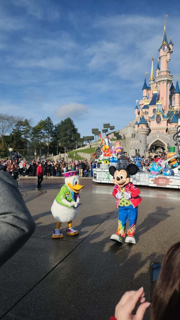 Mickey Mouse et Donald Duck saluent les visiteurs lors d'une parade à Disneyland Paris, avec un château rose et la foule en arrière-plan.