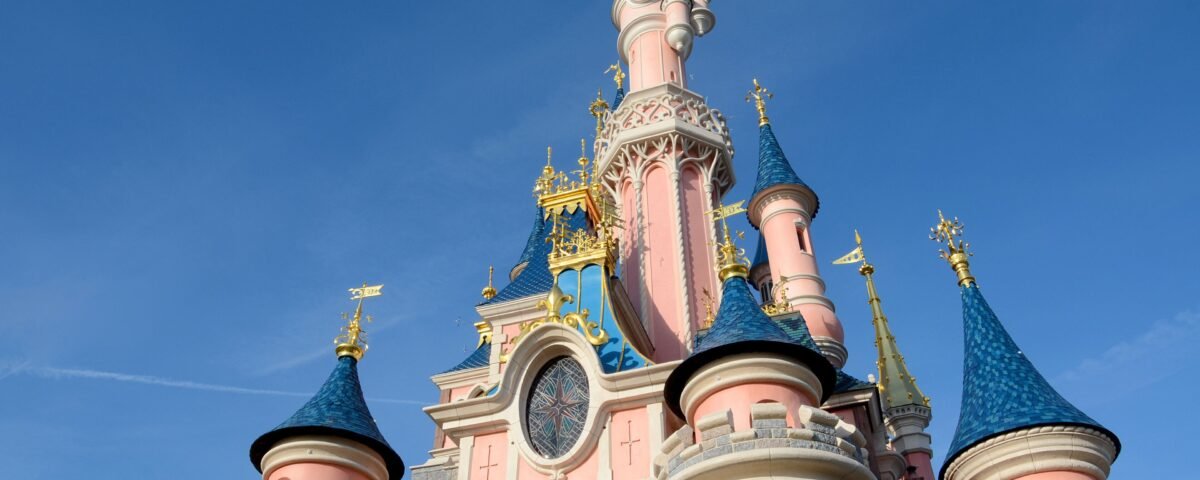 Vue rapprochée d'un château de Disneyland Paris avec des murs roses, des toits bleus en forme de cône et des accents dorés dans un ciel bleu clair.