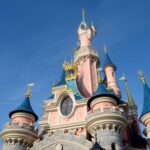 Vue rapprochée d'un château de Disneyland Paris avec des murs roses, des toits bleus en forme de cône et des accents dorés dans un ciel bleu clair.