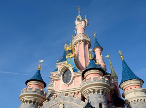 Vue rapprochée d'un château de Disneyland Paris avec des murs roses, des toits bleus en forme de cône et des accents dorés dans un ciel bleu clair.