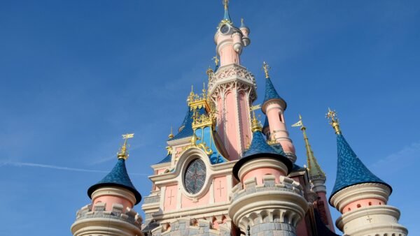 Vue rapprochée d'un château de Disneyland Paris avec des murs roses, des toits bleus en forme de cône et des accents dorés dans un ciel bleu clair.