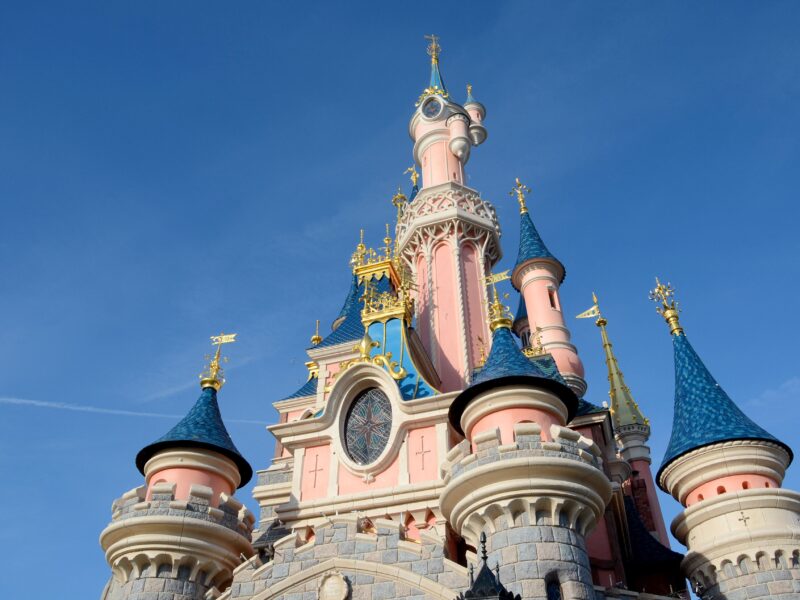 Vue rapprochée d'un château de Disneyland Paris avec des murs roses, des toits bleus en forme de cône et des accents dorés dans un ciel bleu clair.