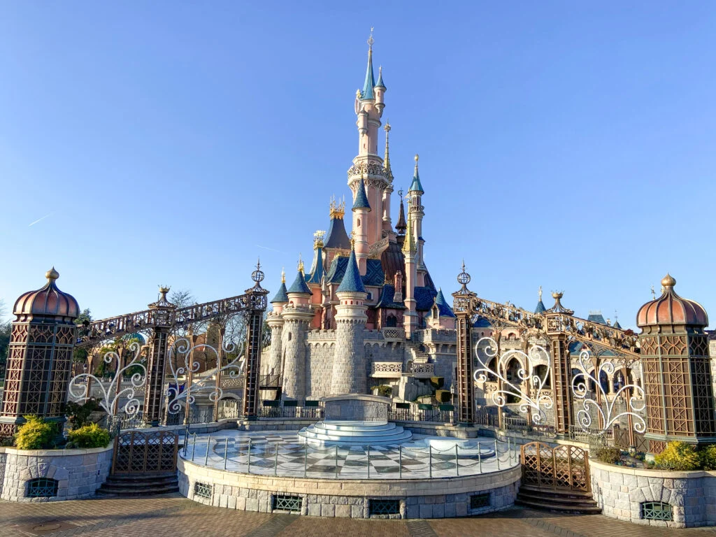 Un château de conte de fées aux tours bleues et roses se dresse derrière des grilles ornées sous un ciel bleu clair à Disneyland Paris, où l'accessibilité et les excellents services aux visiteurs renforcent l'expérience magique du parc à thème.