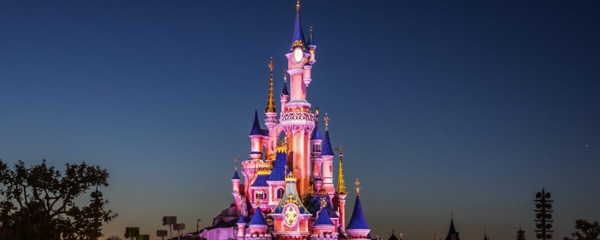 Un château illuminé la nuit sous un ciel clair et sombre, avec des arbres et des silhouettes de tourelles en arrière-plan, évoque l'atmosphère enchanteresse de Disneyland Paris - une source d'inspiration pour tous ceux qui recherchent des conseils avant de visiter.