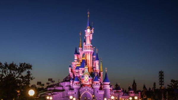Un château illuminé la nuit sous un ciel clair et sombre, avec des arbres et des silhouettes de tourelles en arrière-plan, évoque l'atmosphère enchanteresse de Disneyland Paris - une source d'inspiration pour tous ceux qui recherchent des conseils avant de visiter.