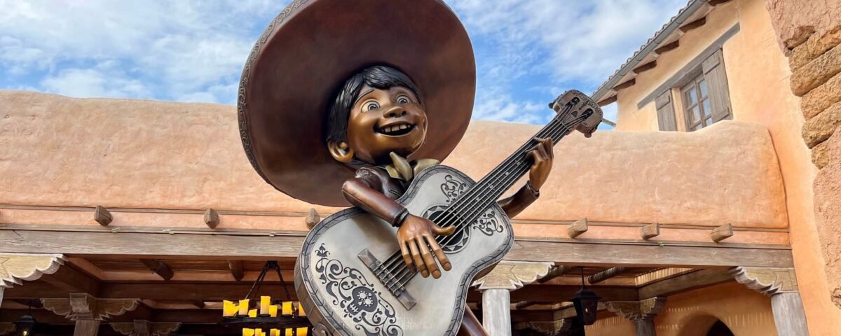 La statue d'un garçon souriant coiffé d'un sombrero et tenant une guitare décorée se dresse devant un bâtiment rustique situé à proximité des boutiques de Disneyland Paris, avec un ciel bleu en arrière-plan.