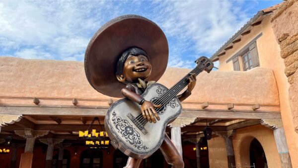 La statue d'un garçon souriant coiffé d'un sombrero et tenant une guitare décorée se dresse devant un bâtiment rustique situé à proximité des boutiques de Disneyland Paris, avec un ciel bleu en arrière-plan.