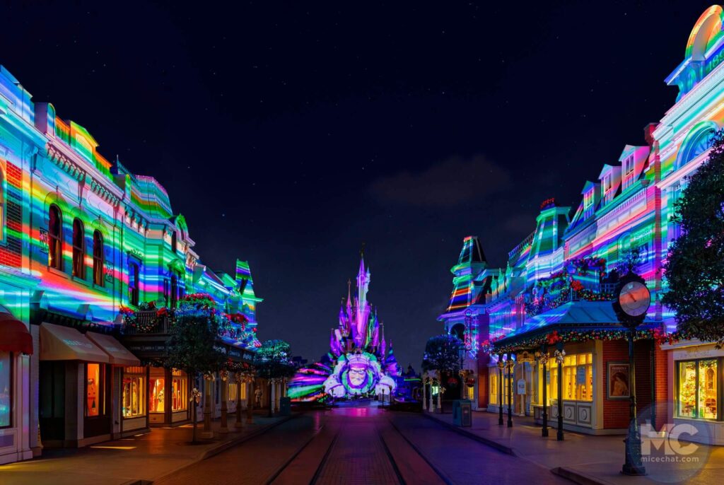 Vue nocturne d'une rue du parc de Disneyland Paris, avec des bâtiments et le château illuminés par des projections lumineuses colorées et rayées à travers différentes zones.