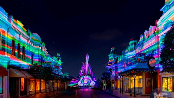 Vue nocturne d'une rue du parc de Disneyland Paris, avec des bâtiments et le château illuminés par des projections lumineuses colorées et rayées à travers différentes zones.