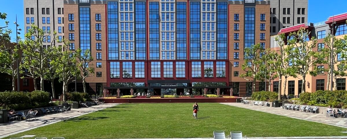 Un grand bâtiment moderne aux hautes baies vitrées, faisant partie d'un Hébergement Disneyland Paris, donne sur une pelouse verte et une cour pavée avec des chaises éparpillées ; une personne seule marche sur l'herbe sous un ciel bleu clair.